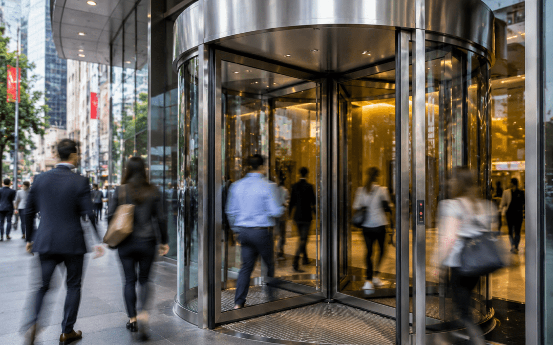 A busy city building entrance with a modern automatic revolving door. Numerous blurred pedestrians in business attire walk in and out, showing high foot traffic, while reflections of the urban environment and warm interior lighting create a dynamic, energetic atmosphere.